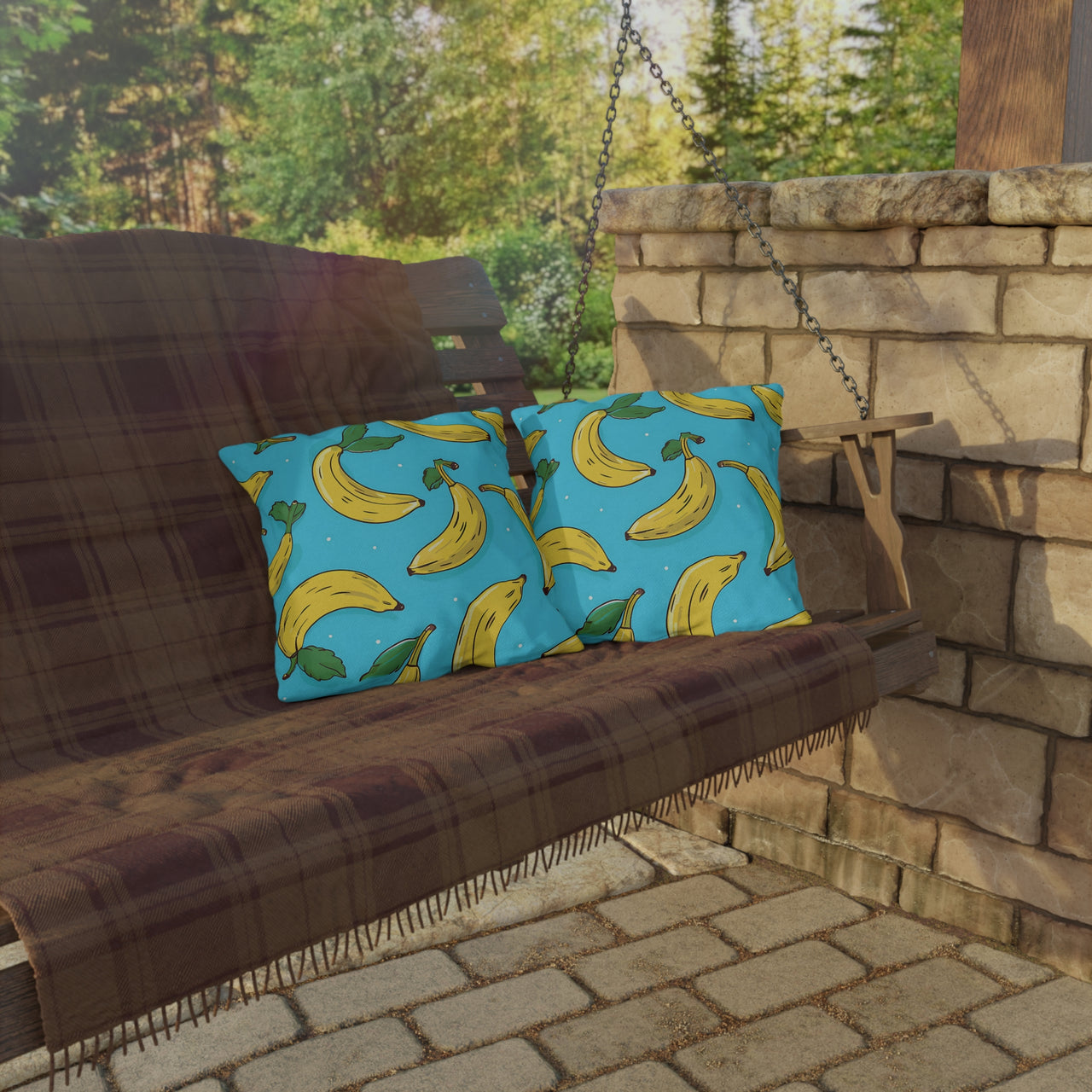 Brown wooden swing with blue pillows featuring banana patterns, set against a stone wall and greenery.